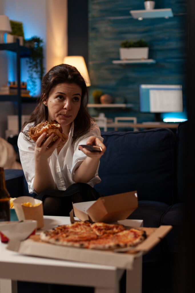 Mulher sentada no sofá comendo hambúrguer delicioso e saboroso segurando controle remoto trocando canais na televisão em frente à mesa com pizza grande. Pessoa navegando pelos canais de TV enquanto come hambúrguer.